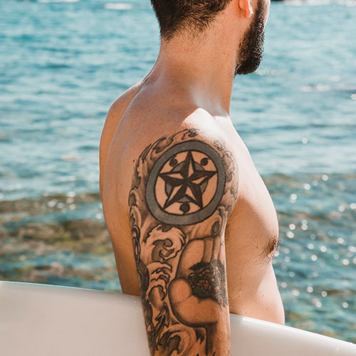 Detalle de un tatuaje ya curado, en un brazo de un chico que mira frente al mar con su tabla de surf en la mano